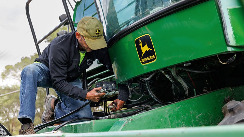 Person wearing a hat and safety glasses while connecting two hoses in front of the cabin of a green machine.